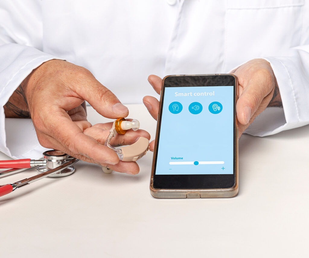 A person in a white coat holds a hearing aid in one hand and a smartphone with a Smart control hearing aid volume app open in the other hand. A red stethoscope lies on the table.
