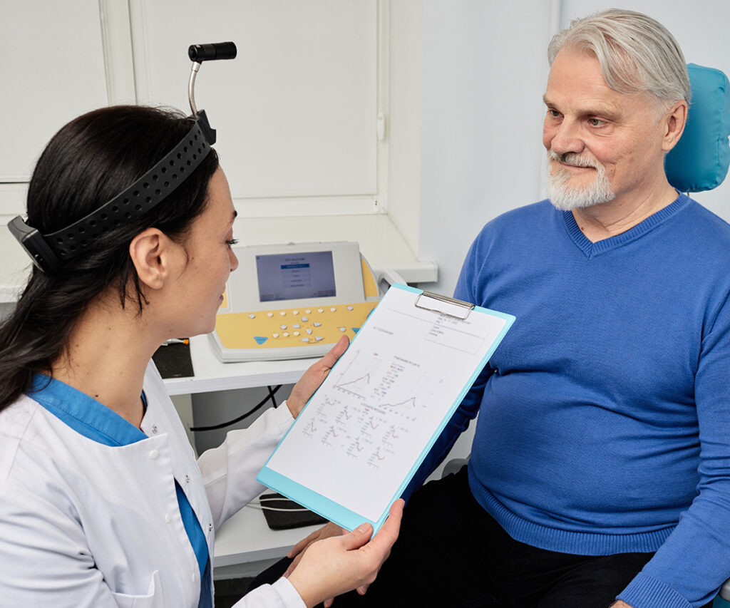 A female doctor wearing a headband shows a medical chart to an older male patient in a blue sweater, who is sitting and smiling in a medical office.
